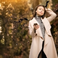 Young woman in a beige coat standing among autumn foliage, listening to music on her smartphone with earbuds.