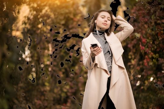 Young woman in a beige coat standing among autumn foliage, listening to music on her smartphone with earbuds.