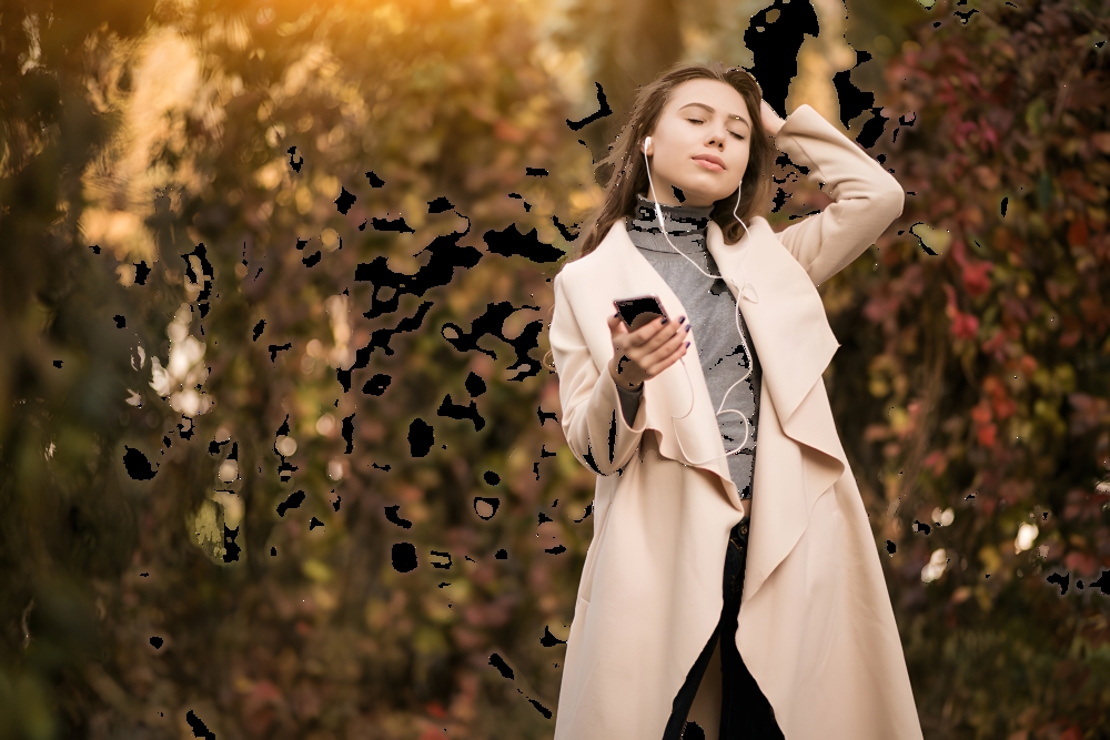 Young woman in a beige coat standing among autumn foliage, listening to music on her smartphone with earbuds.