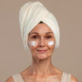 Woman with a towel turban on her head, smiling, wearing white under-eye cream patches on both eyes.