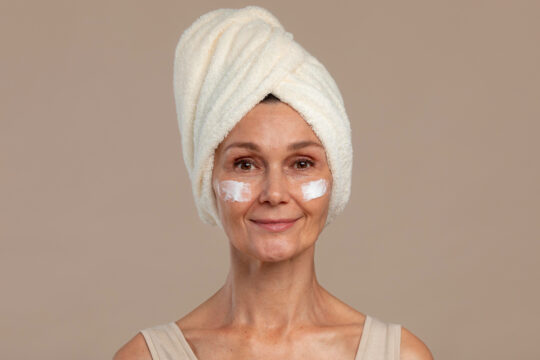 Woman with a towel turban on her head, smiling, wearing white under-eye cream patches on both eyes.