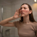 Woman in a beige shirt holds her hand to her ear, listening, in a softly lit bathroom.