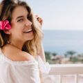 Smiling woman with a pink flower in her hair on a balcony overlooking the sea, wearing a white off-shoulder top.