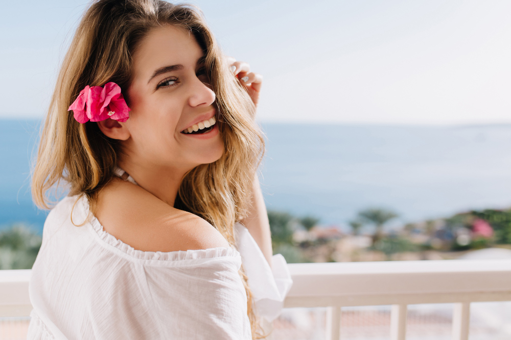 Smiling woman with a pink flower in her hair on a balcony overlooking the sea, wearing a white off-shoulder top.