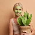 Smiling woman with a green facial mask holding a potted cactus plant against a peach background.