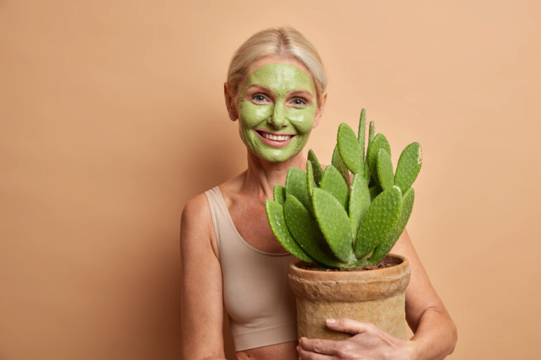 Smiling woman with a green facial mask holding a potted cactus plant against a peach background.