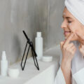 Woman with a towel on her head applies facial cream to her cheeks in front of a small vanity mirror on a counter with skincare bottles.