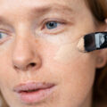 Close-up of a woman applying beige foundation to her cheek with a makeup brush.
