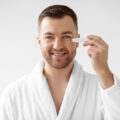 Smiling man in a white bathrobe holds a contact lens applicator to his right eye for insertion.
