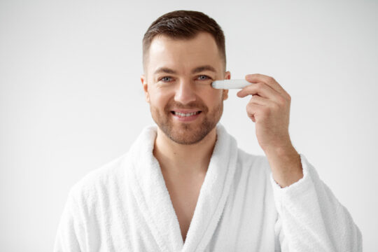 Smiling man in a white bathrobe holds a contact lens applicator to his right eye for insertion.