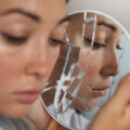 Woman looking at her reflection in a cracked handheld mirror, with a calm, slightly pensive expression.