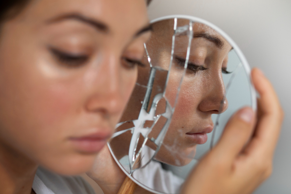 Woman looking at her reflection in a cracked handheld mirror, with a calm, slightly pensive expression.