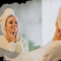 Woman in a white bathrobe with a towel wrapped around her head, smiling at her reflection in a round bathroom mirror.