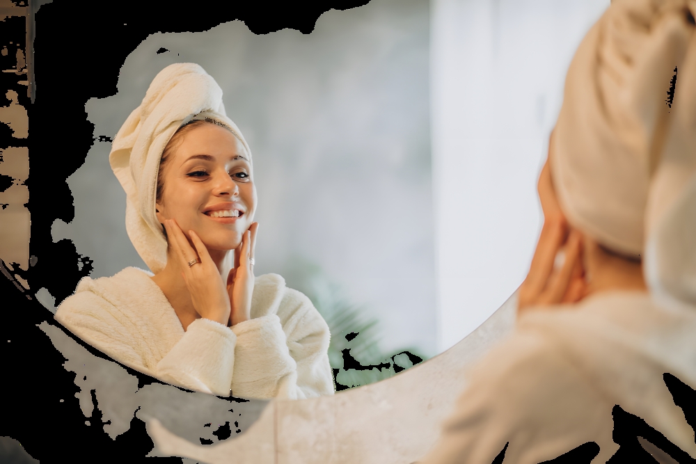Woman in a white bathrobe with a towel wrapped around her head, smiling at her reflection in a round bathroom mirror.