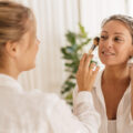 Makeup artist applying makeup with a brush to a smiling woman in a bright, airy room.