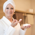 Smiling older woman in a white bathrobe with a towel on her head applies moisturizer using a cotton pad and a cream jar in hand.