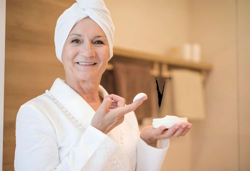 Smiling older woman in a white bathrobe with a towel on her head applies moisturizer using a cotton pad and a cream jar in hand.